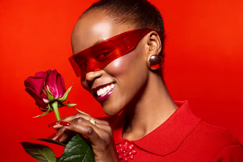 Smiling black woman with dark skin wearing red clothes and futuristic red glasses holds red rose near face on red background. Concept of beauty, fashion, elegance, and love.