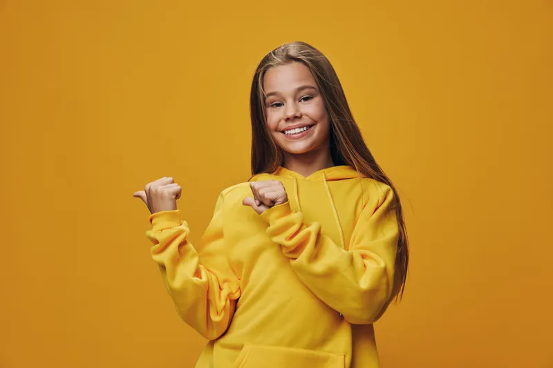 Girl wearing yellow hoodie smiling and pointing with both hands in studio with orange background. Happy child with long hair and casual clothes posing for camera.