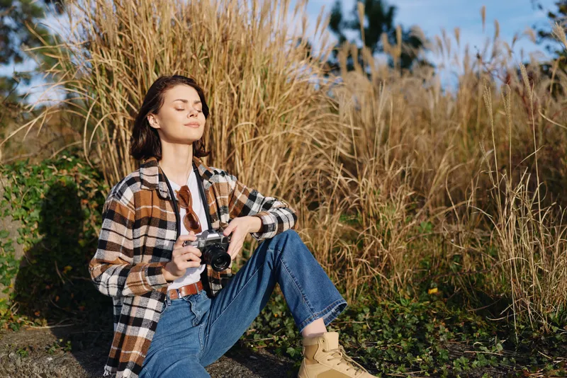 Outdoor portrait of a woman in a plaid shirt and jeans, sitting among tall grass with a camera in hand, enjoying natural light and the peaceful field mood.