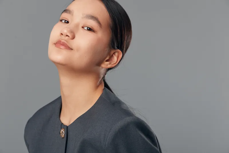 Young Asian woman with smooth skin and tied hair wearing dark collarless jacket posing confidently against gray background in studio portrait lighting.