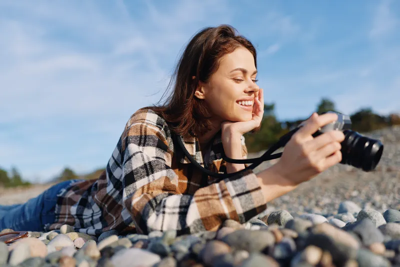 Woman photographer lying on pebbles outdoors, smiling and focused, holding a camera in a casual plaid shirt, enjoying nature and a creative photography moment by the shore