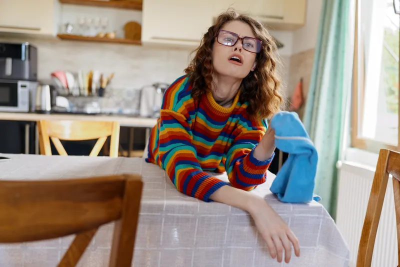 Cheerful woman indoors, wiping a table with a blue cloth, colorful striped sweater, casual lifestyle moment, warm natural light, cozy kitchen background, relaxed posture, bright atmosphere