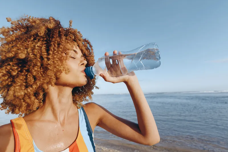 Woman with curly hair drinking water from a plastic bottle by the ocean, summer hydration, outdoor lifestyle, sunny beach atmosphere, focus on thirst relief and wellness, fresh air today