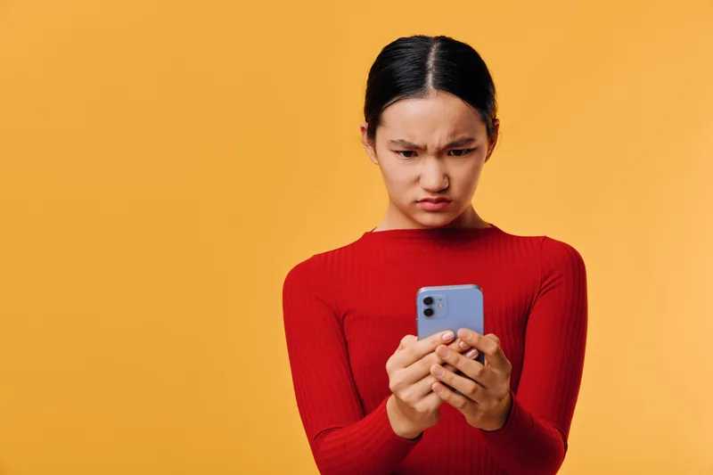Young woman in red sweater looking confused and frustrated at smartphone screen with yellow background. Closeup portrait of displeased female holding mobile device for technology or communication.