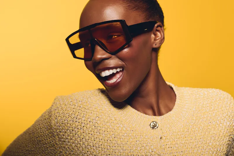 Happy African woman with dark skin wearing oversized sunglasses and yellow sweater smiles broadly against bright yellow background. Concept of fashion, joy, and confident style.