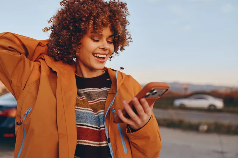 Cheerful woman lifestyle outdoors, smiling with curly hair, using smartphone at sunset, casual jacket, relaxed stance, warm golden light, city road and cars blurred background