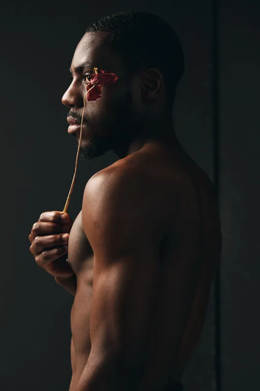 Black man with dark skin holding dried flower near face in creative conceptual visual style. Artistic portrait of African ethnicity with bare torso and moody lighting on dark background.