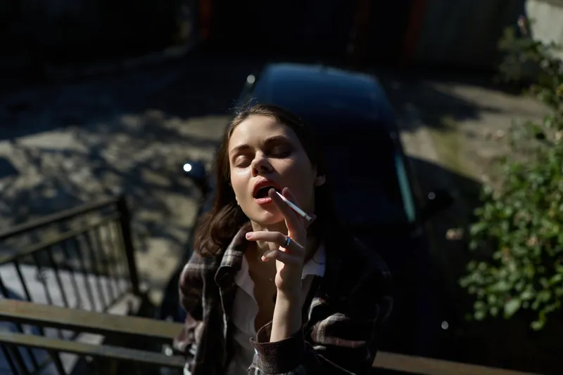 Smiling woman with long dark hair smoking a cigarette on a sunlit balcony. Outdoor lifestyle scene emphasizing casual confidence, modern mood, and everyday routine