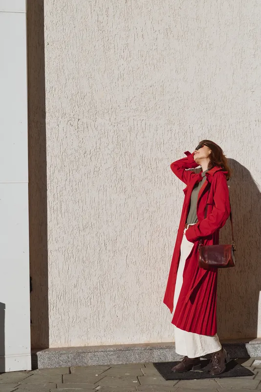 A woman in a bold red longcoat stands against a textured wall, shielding her eyes from the sun while showcasing a confident street fashion moment, casual and poised.