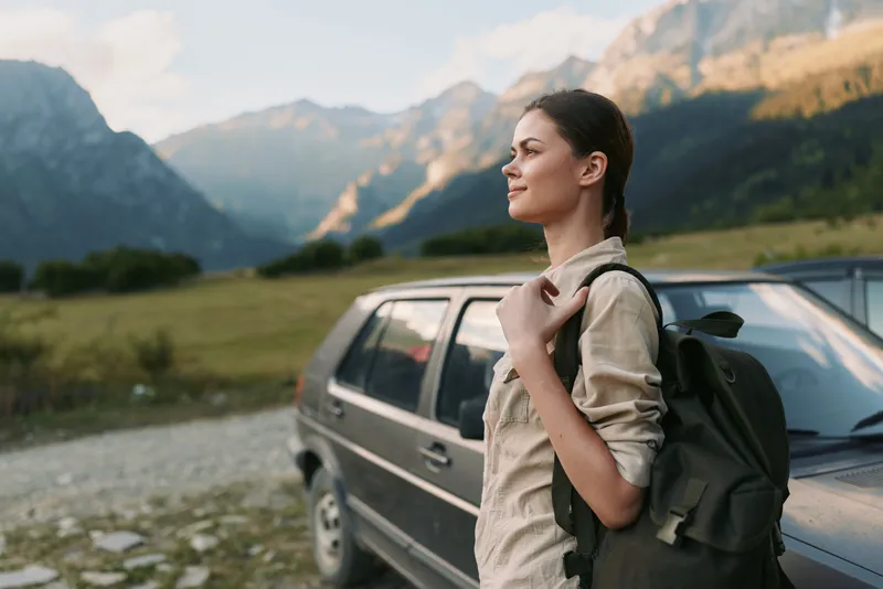 Young woman with a backpack stands near a parked car, gazing at dramatic mountains, enjoying a peaceful outdoor moment with sunlight on peaks.