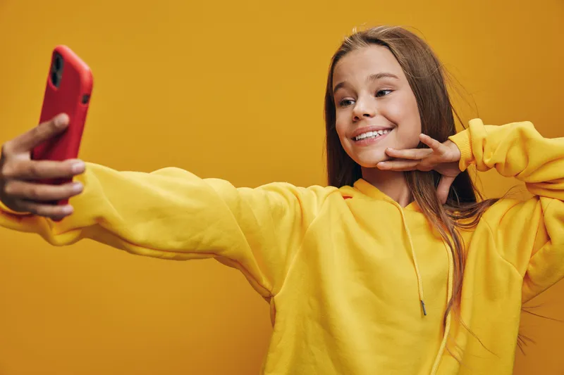 Girl in yellow hoodie taking selfie with red smartphone on yellow background smiling and posing confident for social media portrait and casual lifestyle shot.