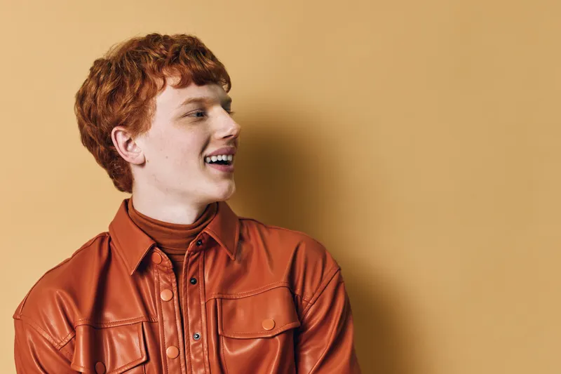 Young man with red hair smiling and looking sideways wearing a brown leather jacket and turtleneck sweater against beige background in studio portrait.