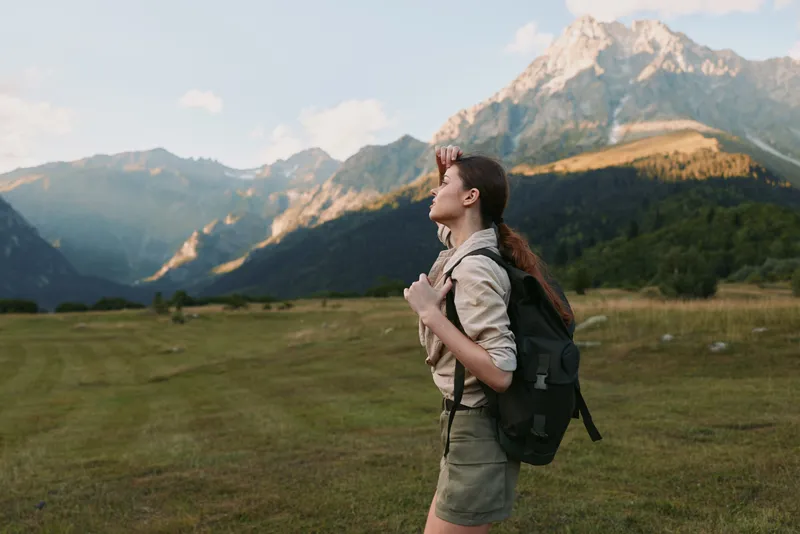 Young woman with a backpack hikes across a grassy valley toward distant mountains, captured in soft light that highlights exploration, independence and a deep connection with nature.