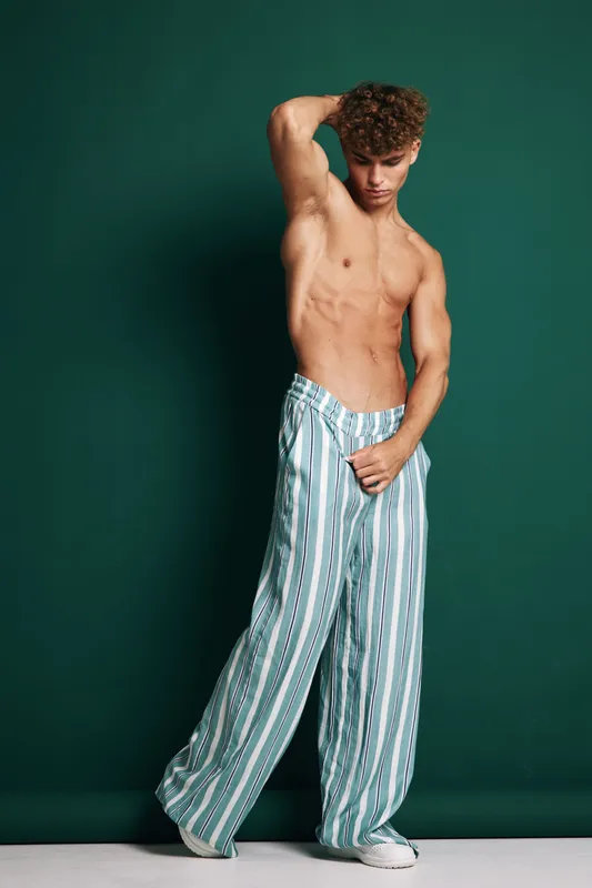 Young muscular man posing barechested in loose striped pants against green background, showing body expression and fashion style with casual white shoes on studio floor.