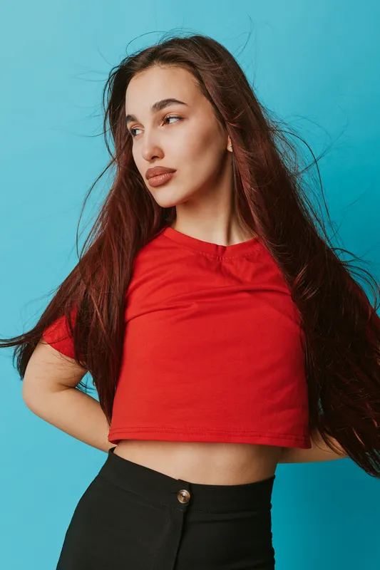 Young woman with long hair wearing red crop top and black pants posing against blue background looking sideways with confident expression in studio portrait fashion concept.