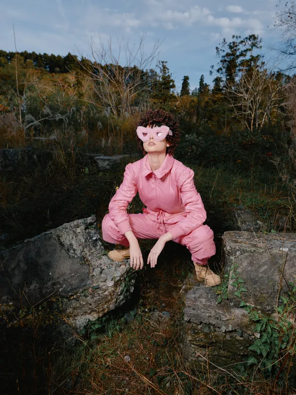 Person wearing pink jumpsuit and mask sits on a rock in a forested area, creating a surreal mood of quiet solitude amid autumn trees and wild greenery.