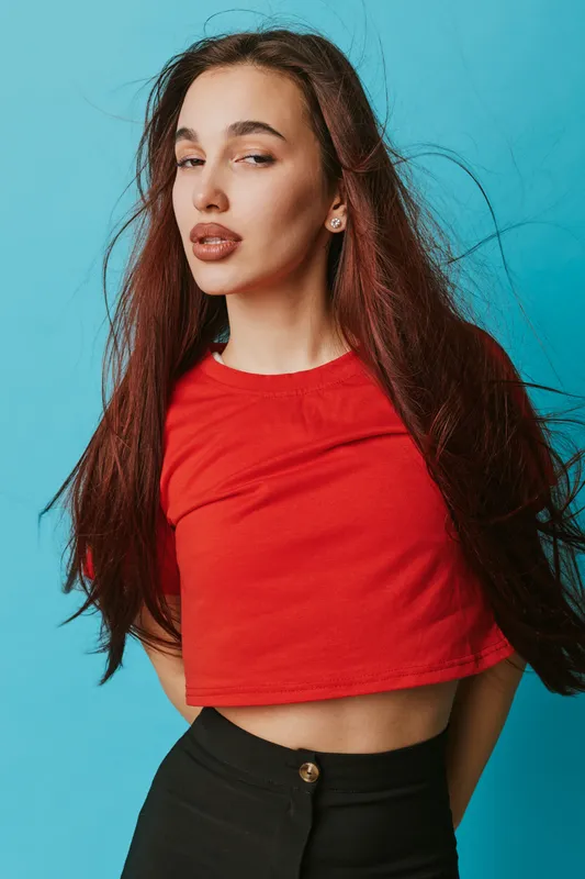 Young woman with long brown hair wearing red crop top and black pants posing against blue background. Stylish female model with confident expression and casual fashion look.
