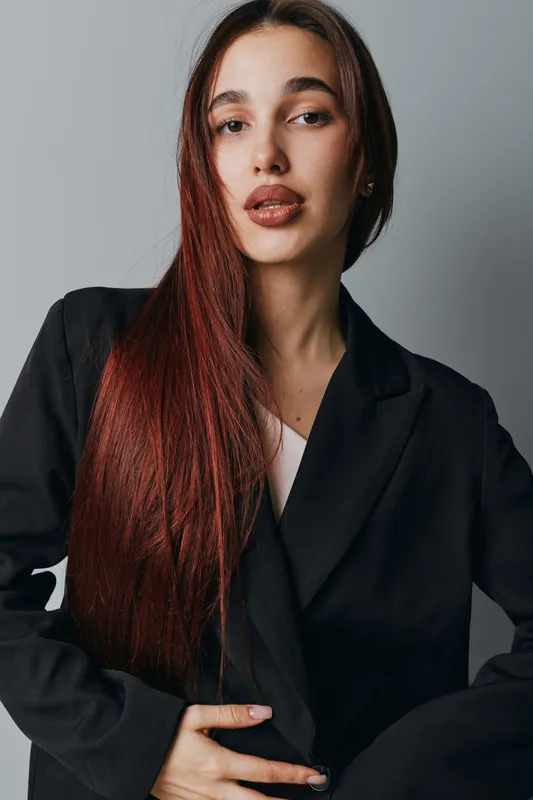 Woman with long red hair and natural makeup wearing black blazer posing with hand on waist against gray background. Fashion portrait of confident female model in studio lighting.