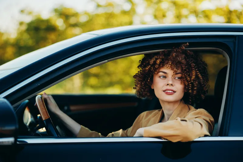 Young woman drives a car, casual chic style, curly hair, retro film colors, smiling expression, everyday lifestyle shot, steering wheel visible, outdoor daylight, soft golden hues, candid travel