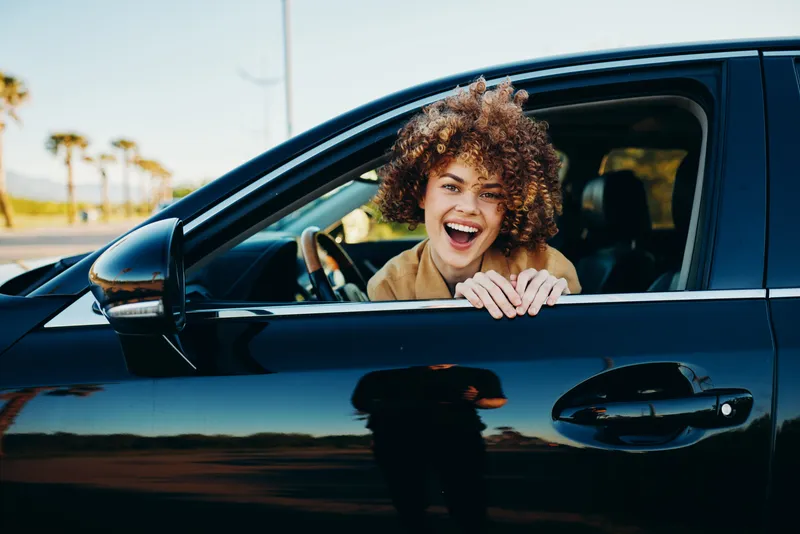 A joyful woman sits in a moving car with the window down, sun on her smile. A candid lifestyle shot with retro film color evokes freedom and everyday happiness.