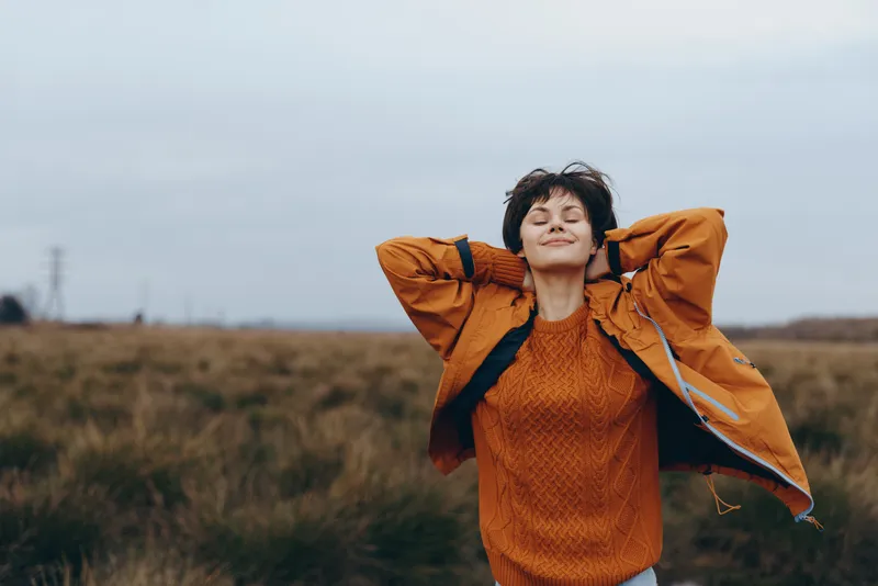Woman lifestyle outdoors in a grassy field, rust jacket, hands on head, windy cloudy sky, natural light, candid portrait, relaxed smile