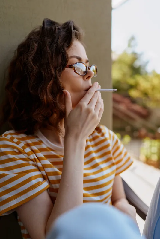 A woman with curly hair wearing a striped shirt sits by a window, smoking a cigarette in a relaxed home setting. Casual portrait conveying mood, comfort, and daily self care and self love.