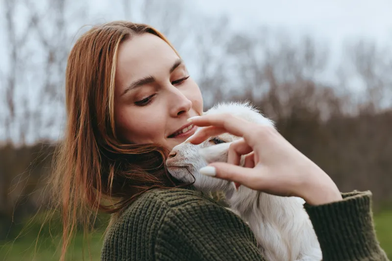 Young woman with long brown hair gently holds and cuddles a white baby goat outdoors during a calm autumn day, expressing warmth and connection in a natural lifestyle setting.