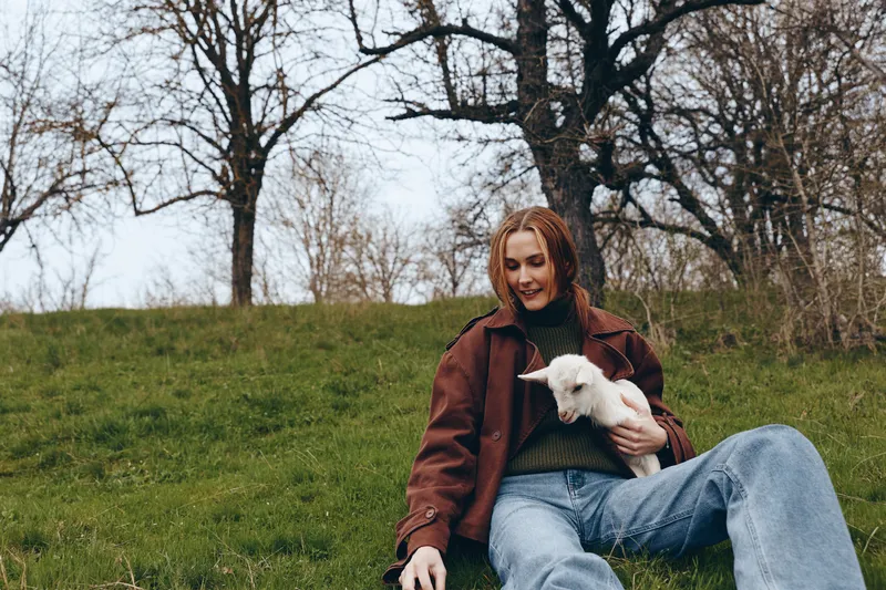 Smiling woman sitting on grass outdoors, cradling a white goat kid. Casual brown jacket and denim. Peaceful countryside scene conveying animal companionship, relaxed lifestyle, and gentle wellbeing.