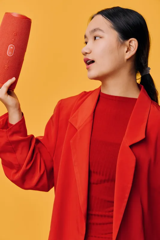 Young woman holding portable speaker in hand, wearing red jacket and looking at device with interest against yellow background. Technology, entertainment, music and casual style.