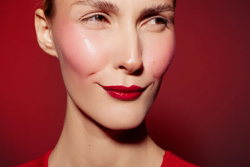 Close-up of a woman with red lipstick and blush on her cheeks against a red background. The image highlights makeup, beauty, and facial expression with soft lighting.