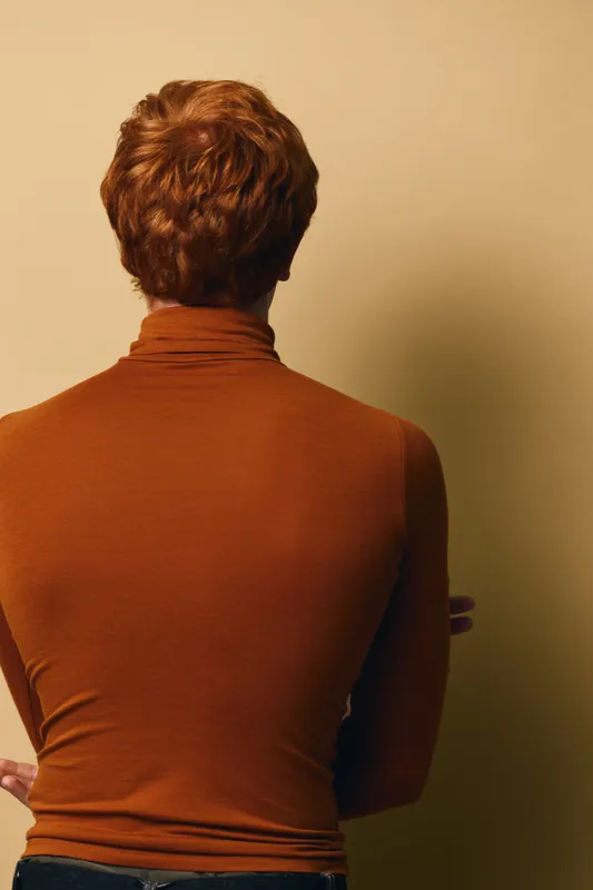 Person with short curly hair wearing brown turtleneck sweater stands with back to camera against beige background indoors.