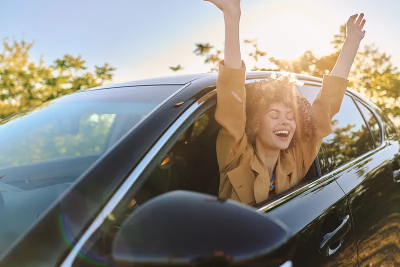 A joyful woman rides in a car with her arms raised in celebration, captured in warm retro film tones that evoke carefree road trip energy and sunlight. Her hair curls around her shoulders, and the