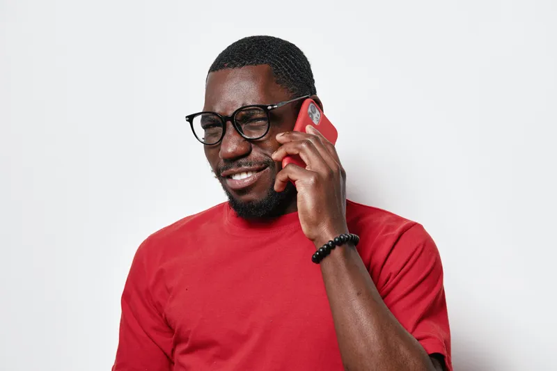 African American man with dark skin wearing glasses and red shirt talking on smartphone with smile against white background. Casual communication and technology concept.