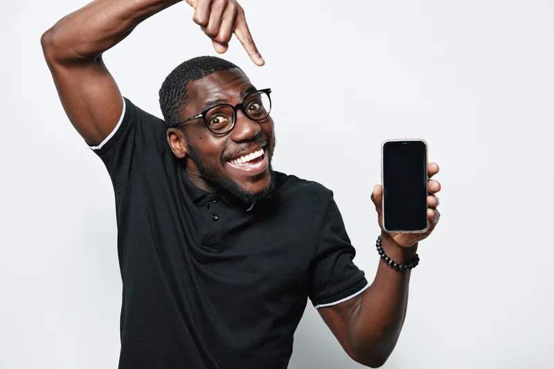African American man with dark skin and glasses smiling and pointing at smartphone screen in black polo shirt on white background. Happy male showing mobile device for technology.