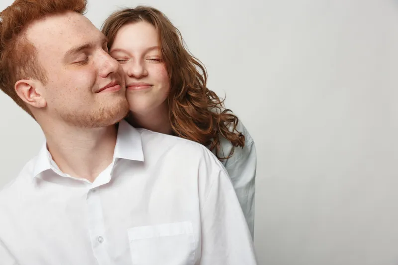 Happy young couple with closed eyes embraces gently, showing love and affection. Casual clothing and neutral background create warm and intimate mood of togetherness and trust.