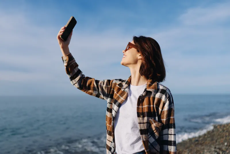 Selfie, woman and smartphone by the seaside with clear sky and calm water, casual plaid shirt, portrait moment outdoors for lifestyle, travel, beach day and personal post