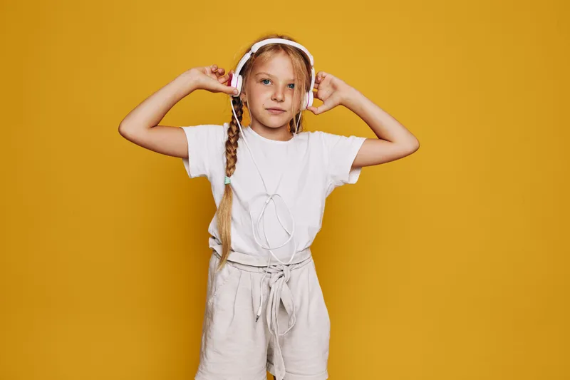 Young girl with headphones listens to music and adjusts them on her head. She stands in front of a yellow background wearing casual clothes.
