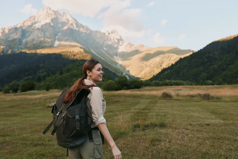 A young woman with a backpack enjoys hiking in the mountains on a sunny day, surrounded by green fields and clear blue sky. Lifestyle concept of outdoor adventure.