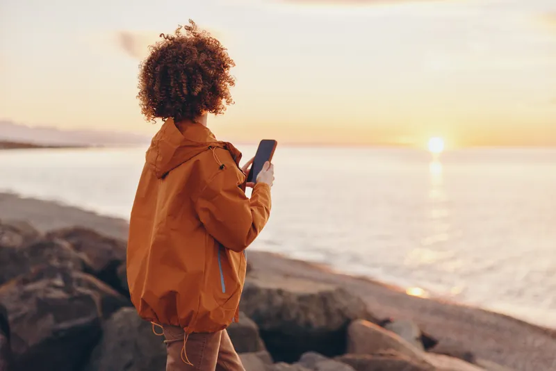 Woman outdoors at seaside during sunset, curly hair, warm jacket, holding a smartphone, lifestyle moment on rocky shore, golden sunlit ocean horizon, soft evening breeze, quietly smiling