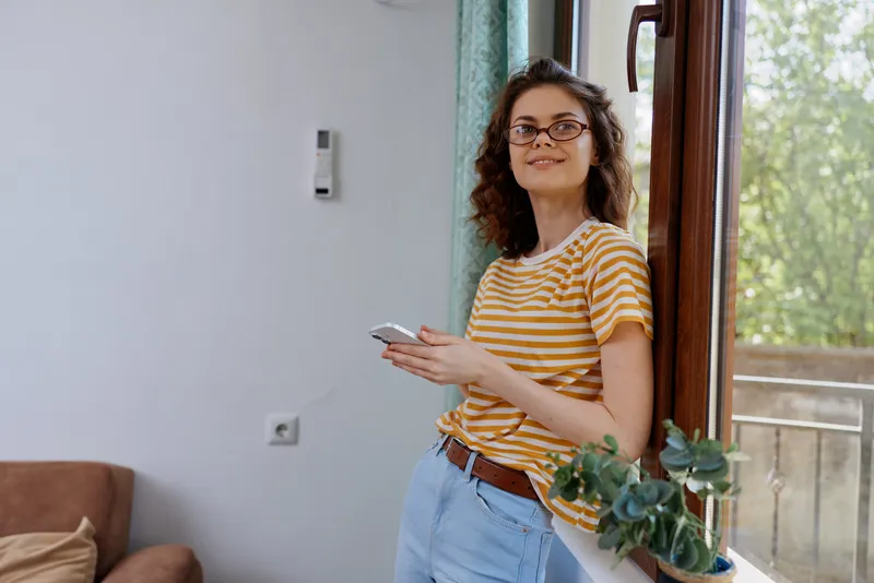 A young adult woman stands by a doorway indoors, wearing a striped tshirt and jeans, looking at her phone with a relaxed smile in a sunlit home interior beside a window