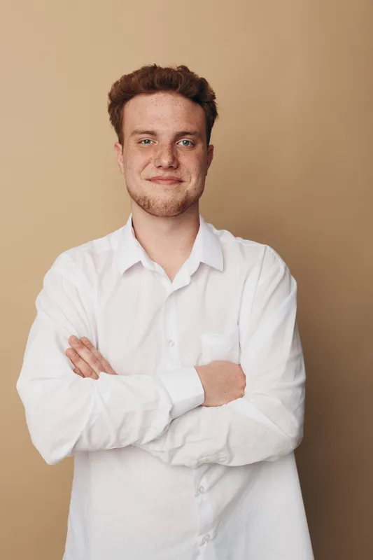 Confident young man with red hair and beard wearing white shirt stands with arms crossed against beige background. Portrait of casual style, positive mood, and natural lighting.