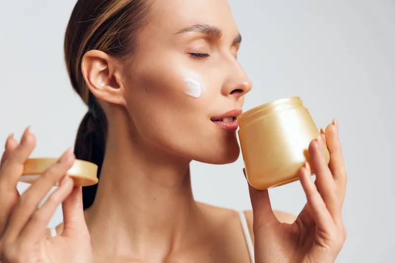 Young woman with closed eyes applying cream on cheek while holding jar near face. Concept of skincare, beauty routine, and facial care with soft natural lighting on white background.
