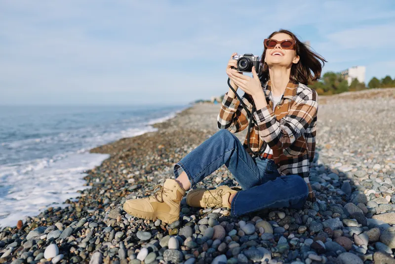 Woman photographer sitting on a pebble beach, wearing plaid shirt and boots, capturing the sea landscape with a camera under a sunny sky, enjoying outdoor calm.