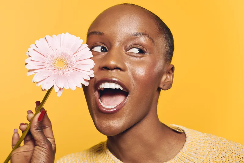 Happy African woman with dark skin holds pink flower near face smiling, isolated on yellow background. Concept of beauty, joy, nature, and positive emotions in studio portrait.