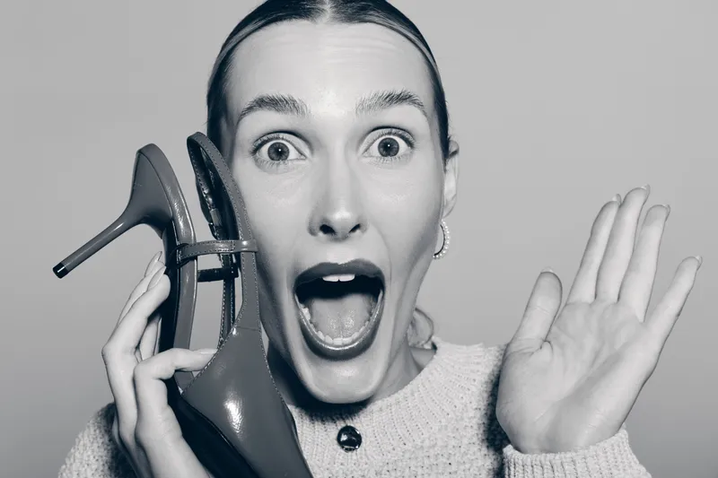 Caucasian woman with light skin holds high heel shoe near ear as if talking, mouth open in surprise, wearing sweater and earrings, isolated on grey background.