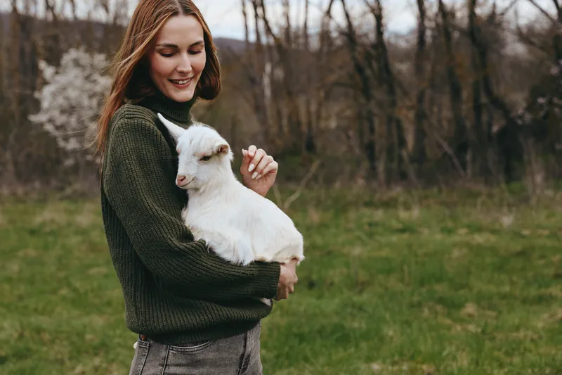 Young woman in a green sweater holds a white baby goat outdoors in a grassy field during early spring with a gentle smile on her face and bare trees in the background