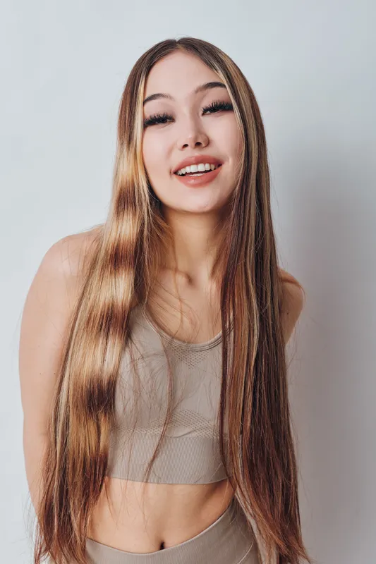 Young woman with long straight hair and casual outfit smiling at camera in studio. Female model with natural makeup and beige top posing for portrait shot with confident expression.
