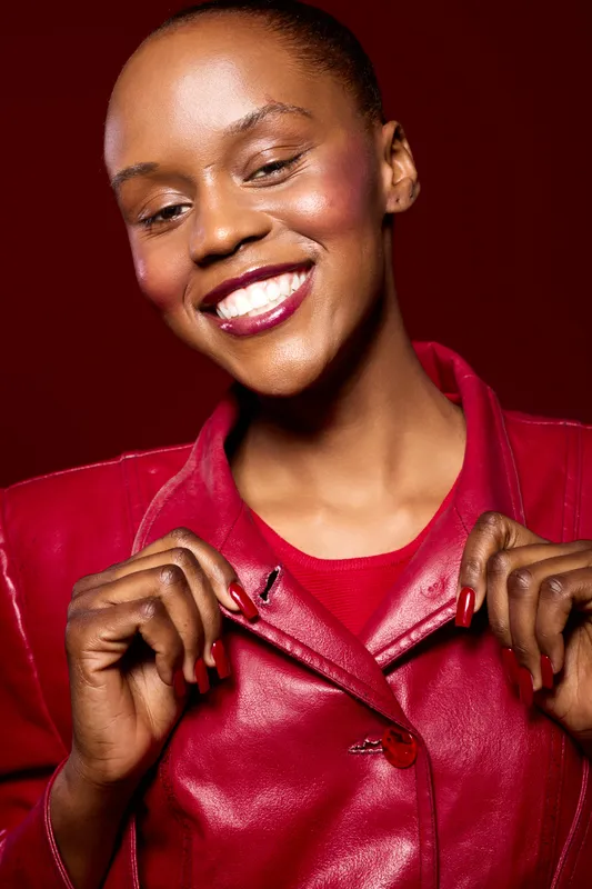 Happy African woman with dark skin wears red leather jacket, smiles brightly against red background. Concept of fashion, confidence, beauty, and positive emotions in studio portrait.
