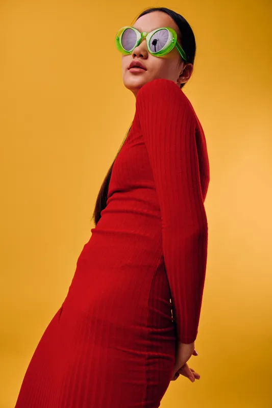 Woman wearing red dress and green sunglasses posing against yellow background. Fashion model with long hair in stylish outfit looking confident and modern in studio light.