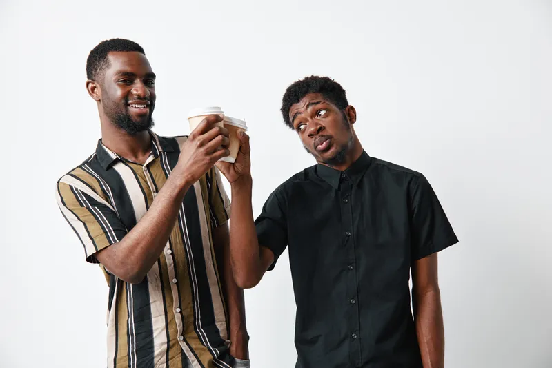 Two young black men with dark skin clinking coffee cups in celebration, casual clothing and friendly expression on white background for friendship and joy.